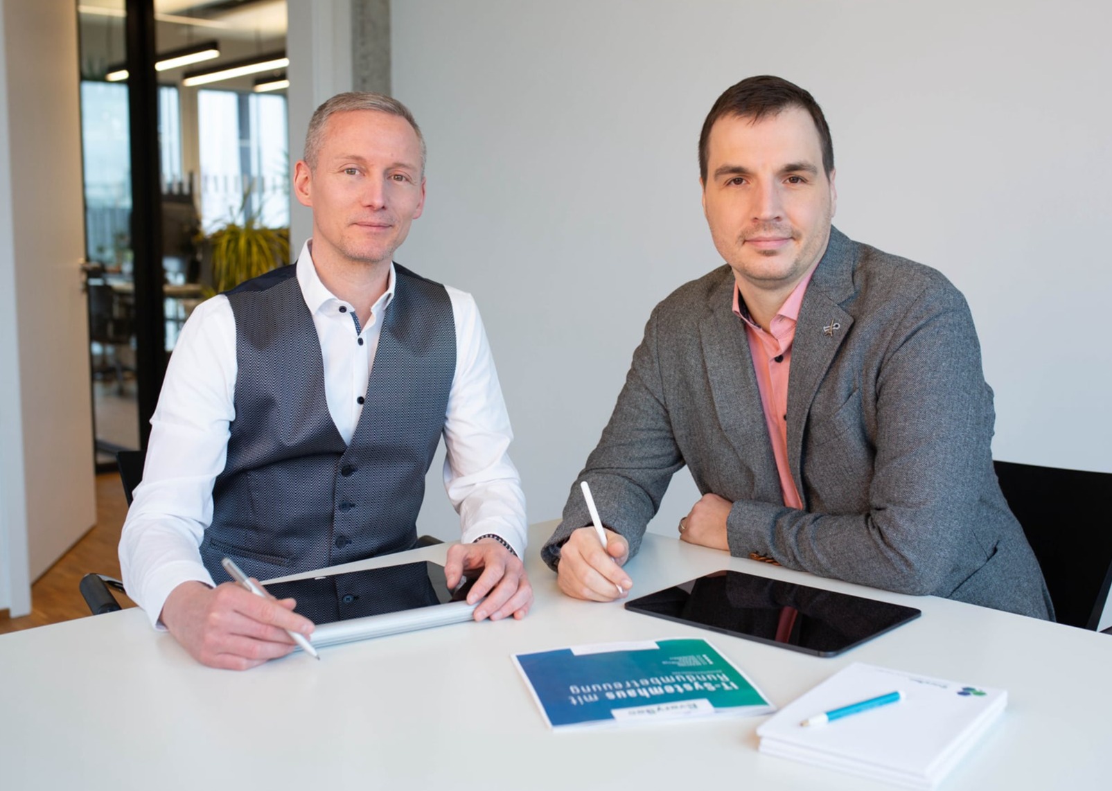 Geschäftsführer Adrian Barthel und Prokurist Matthias Hümmer sitzen nebeneinander an einem Konferenztisch und haben Tablets vor sich.