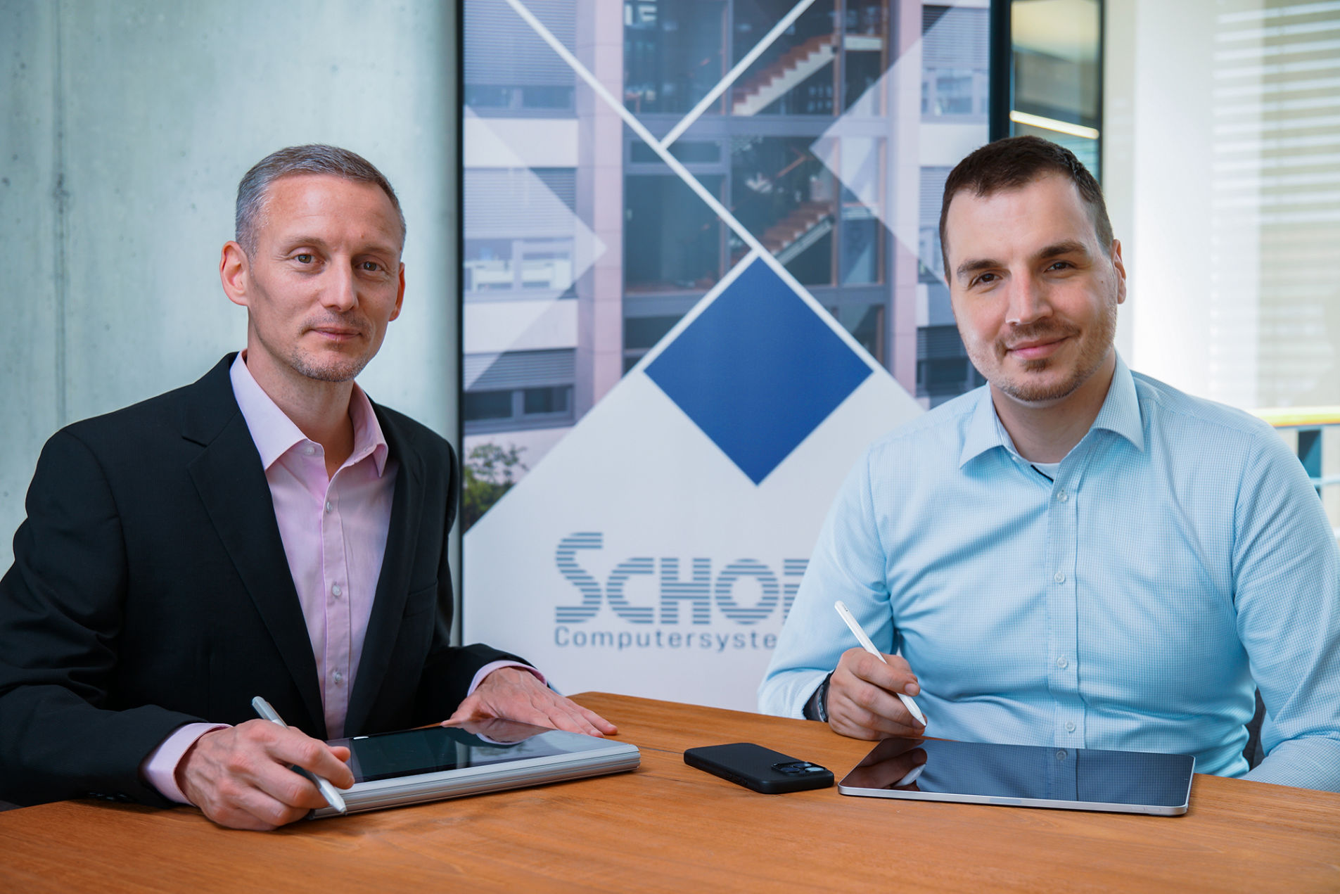 Geschäftsführer Adrian Barthel und Prokurist Matthias Hümmer sitzen nebeneinander an einem Konferenztisch und haben Tablets vor sich.